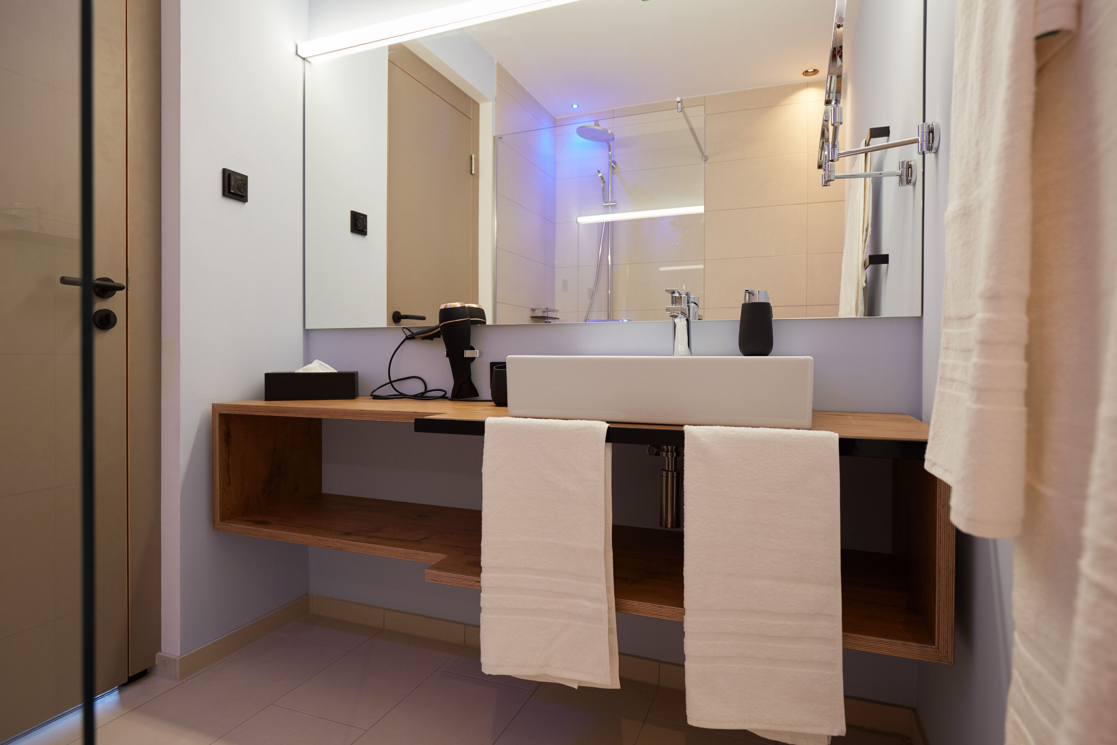 Une salle de bain moderne avec un grand miroir, un lavabo et une douche. Des serviettes sont accrochées au mur, et il y a aussi un porte-serviettes.