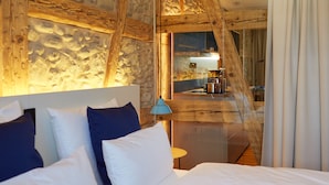 Un dormitorio con pared de piedra, vigas de madera, cama con ropa de cama blanca y azul, mesita de noche y vistas a la cocina.