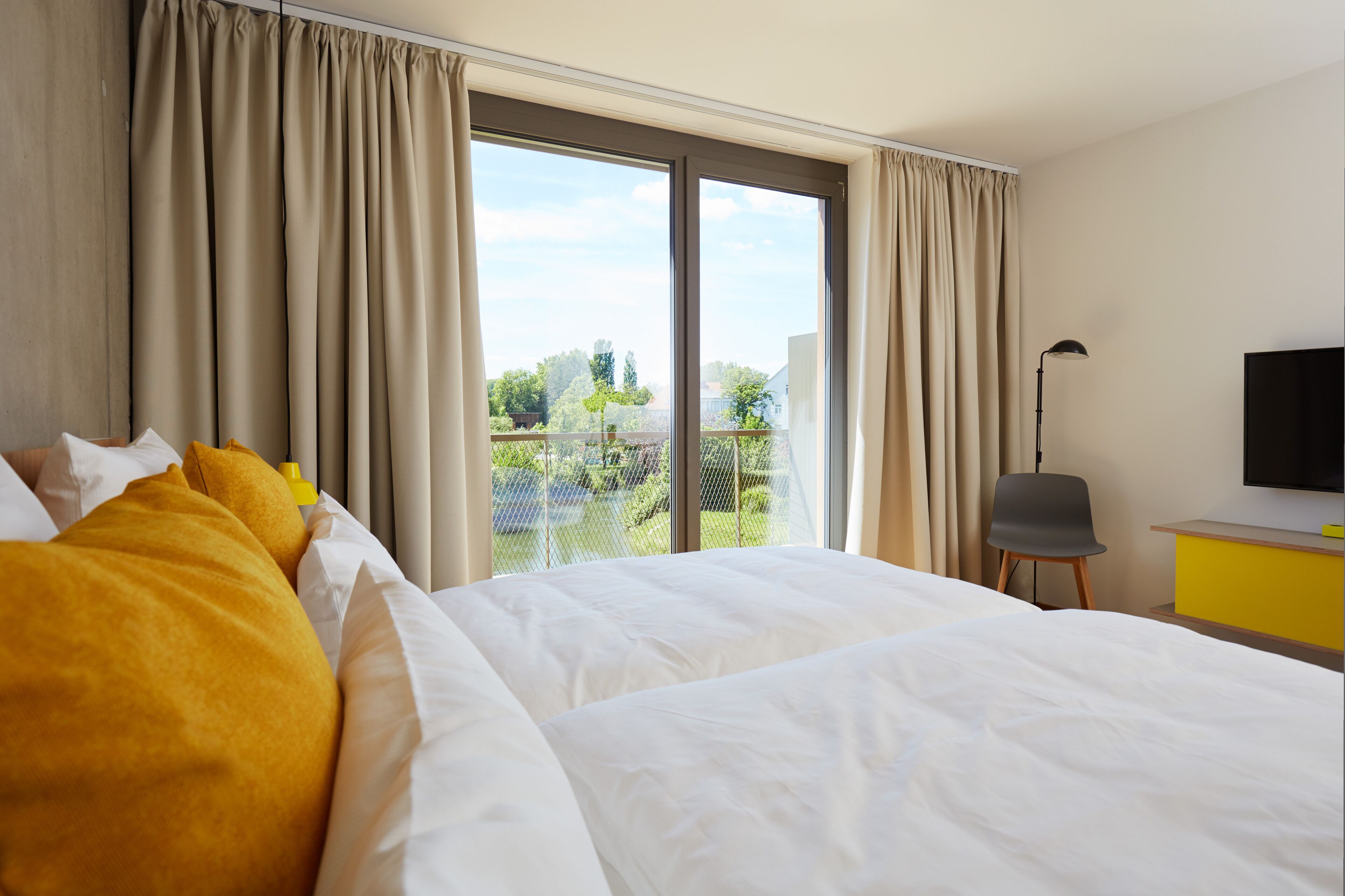 A hotel room with a large bed, a desk, a chair, a TV, and a balcony with curtains.