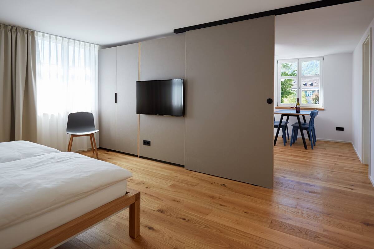 Ett modernt hotellrum med en säng, en TV som är monterad på väggen, en matplats med ett bord och stolar samt ett stort fönster med utsikt över grönområden.
