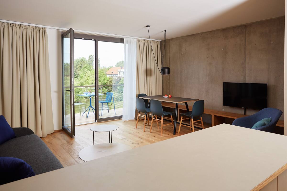Ett modernt vardagsrum med en soffa, ett matbord med stolar, en tv och en skjutdörr i glas som leder ut till ett utomhusområde.