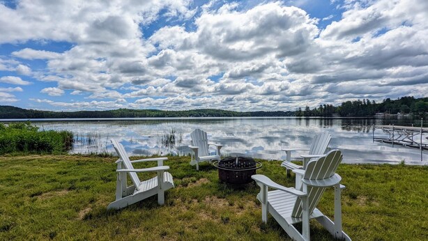 A lakeside view with three Adirondack chairs, a fire pit, and a dock extending into the water.