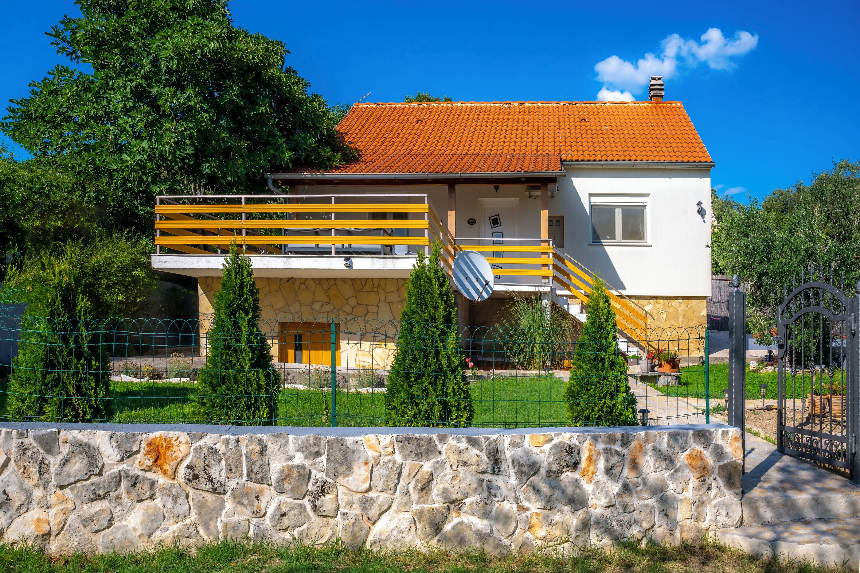 Une maison à deux étages, avec des fondations en pierre, un toit recouvert de tuiles rouges et une balustrade jaune à l’étage supérieur.