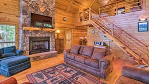 Un interior de cabaña acogedor con chimenea de piedra, paredes de madera, un sofá grande y una escalera que conduce a un nivel superior.