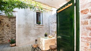 Un petit espace extérieur aménagé avec une table et deux tabourets, une fenêtre ornée de fleurs et une porte verte.