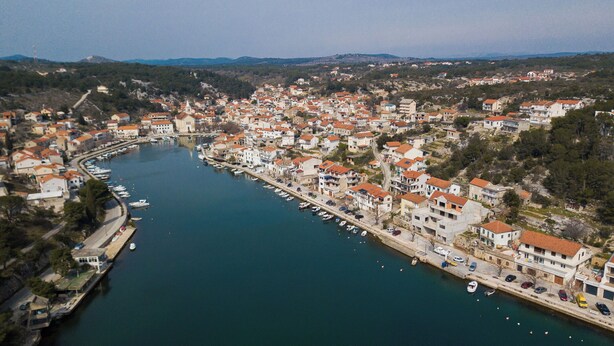Vue aérienne d’une ville côtière avec un port de plaisance, des bâtiments aux toits recouverts de tuiles rouges et un ciel dégagé.