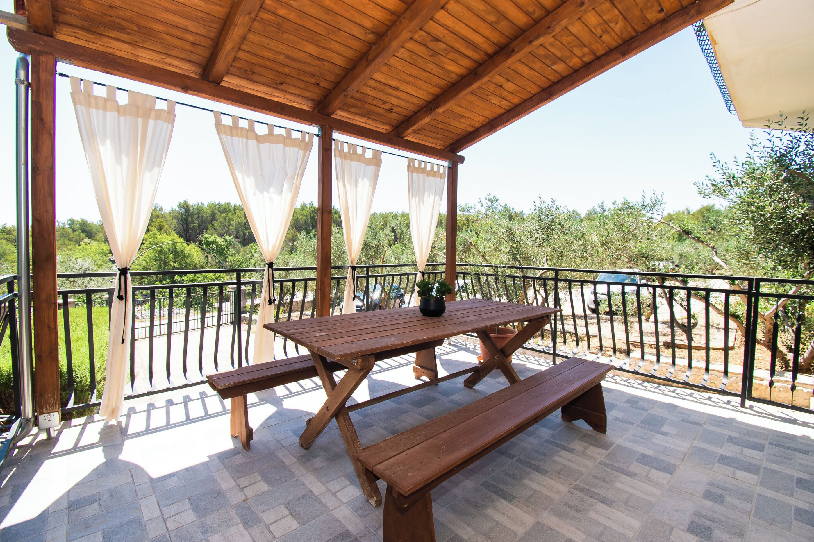 Une terrasse extérieure en bois, dotée d’une table de pique-nique et de bancs, entourée d’une rambarde en métal et de rideaux légers.