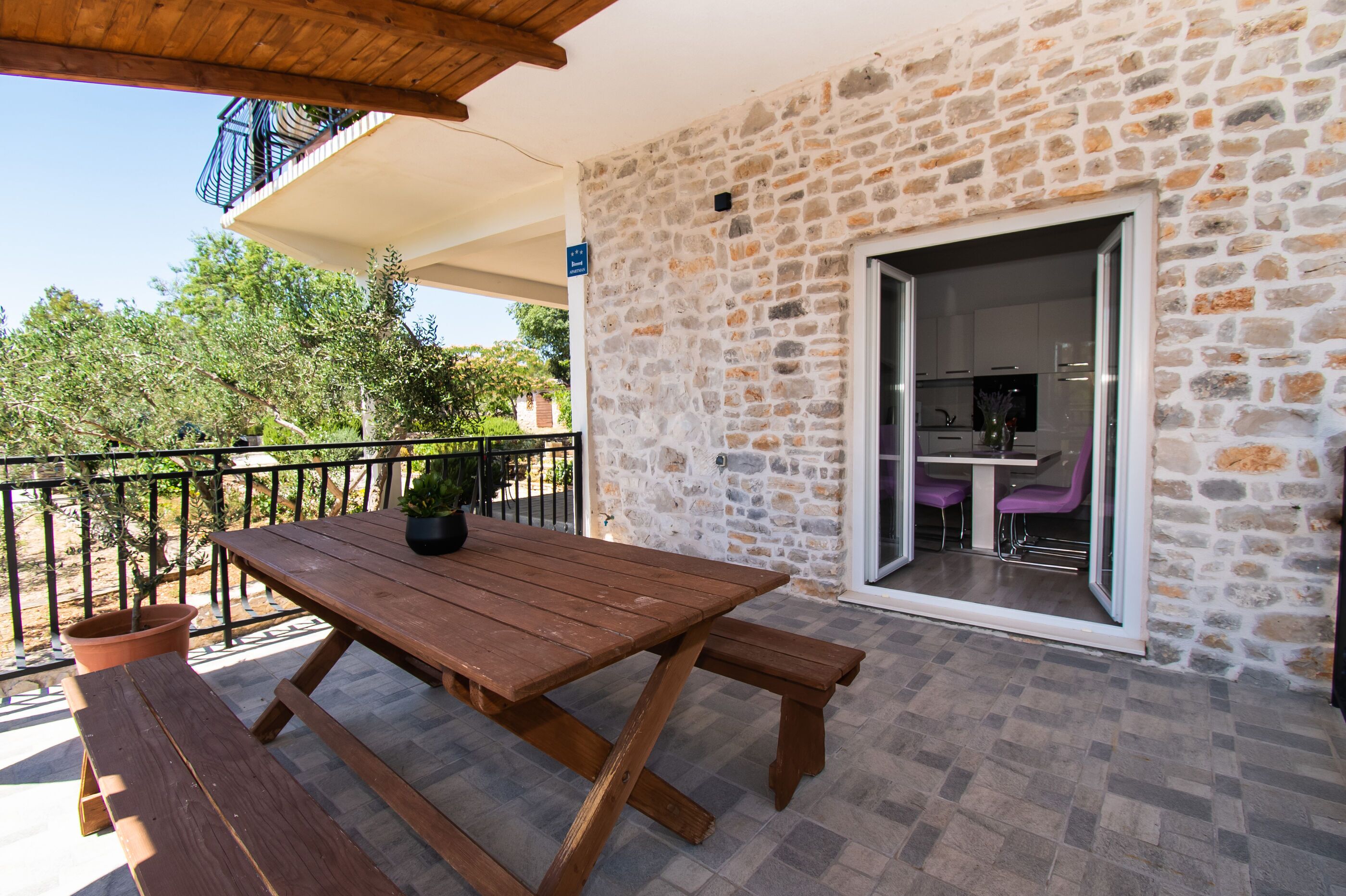 A wooden table with benches on a balcony with stone walls, a potted plant, and a view of a kitchen through a glass door.