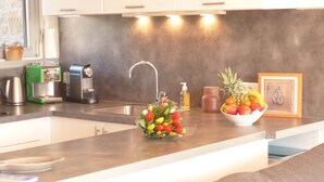 A modern kitchen with a stainless steel sink, a coffee maker, a fruit bowl, and a framed picture on the counter.