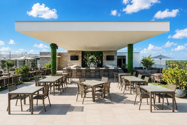 Un patio exterior con varias mesas y sillas, una zona de bar y vistas a la ciudad.