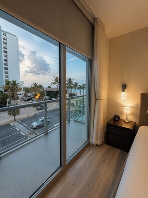 Quarto de hotel com uma janela grande, mesa de cabeceira com luminária e vista para a rua e palmeiras.