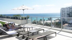 A rooftop terrace with sun loungers, a table, and an umbrella, overlooking a coastal view with buildings and palm trees.