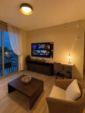 A modern living room with a flat-screen TV, a sofa, a coffee table, and a desk with a lamp. - 1BR Gorgeous Parcial view Beach, Resort 4 Pax (Hollywood)