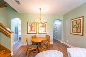 A dining area with a round table, four chairs, and a chandelier. There are framed pictures on the walls and a staircase to the left.