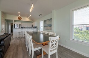 A spacious dining area with a long table and white chairs, a kitchen with white cabinets, and a large window with blinds. - Savannah - Nags Head MP 16, Oceanfront, Direct Beach Access, Pool, Hot Tub, Pool Table (Nags Head)