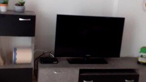 A living room with a flat-screen TV on a black entertainment unit, a small table with a plant, and a white air conditioning unit on the wall. - Immaculate 2-bed Apartment (Paiania)