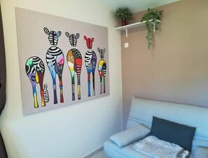 A room with a light blue sofa, a wall-mounted artwork of colorful abstract shapes, and a small plant on a shelf.