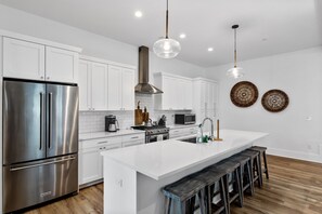 A modern kitchen with white cabinetry, stainless steel appliances, a large island with stools, and decorative wall art. - Stay @ Mimosa Manor - Private Pool, Steps to Beach (Folly Beach)