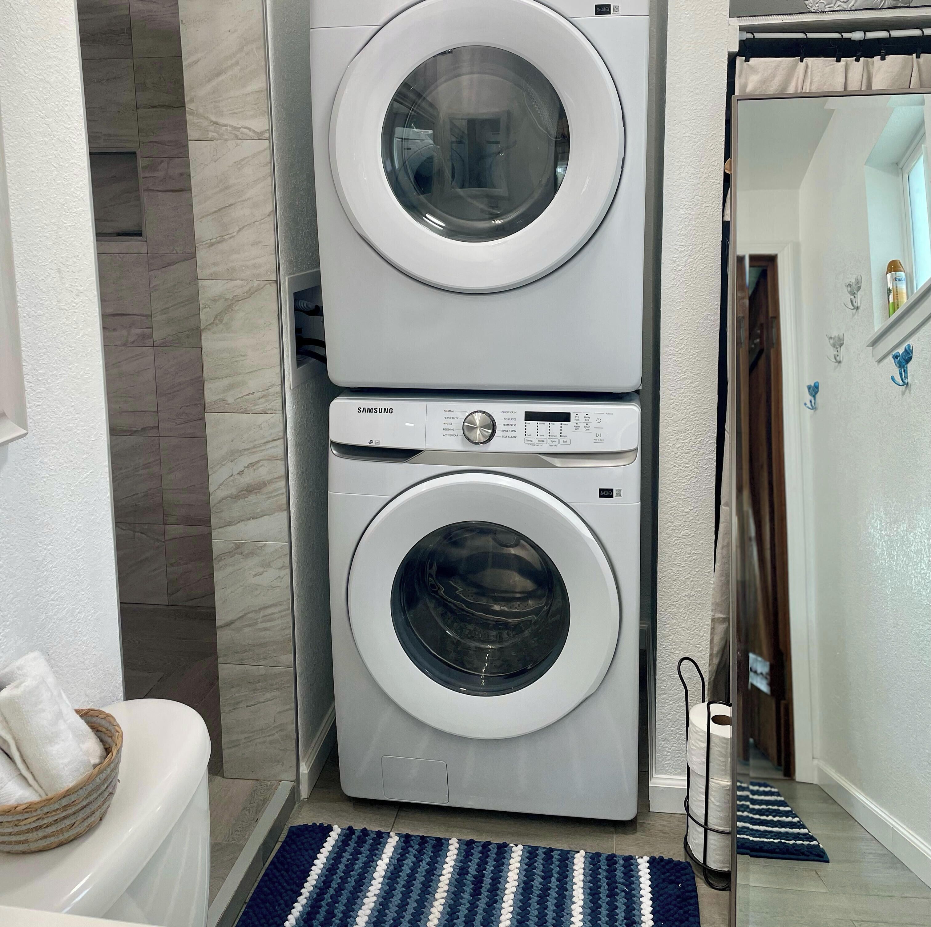 Una lavadora y secadora apiladas en un baño con piso de baldosas, un tapete a rayas y un cesto para toallas.