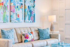 Uma sala de estar moderna com um sofá bege, almofadas coloridas, arte abstrata na parede, uma luminária em uma mesa lateral e um tabuleiro azul com um enfeite de estrela.