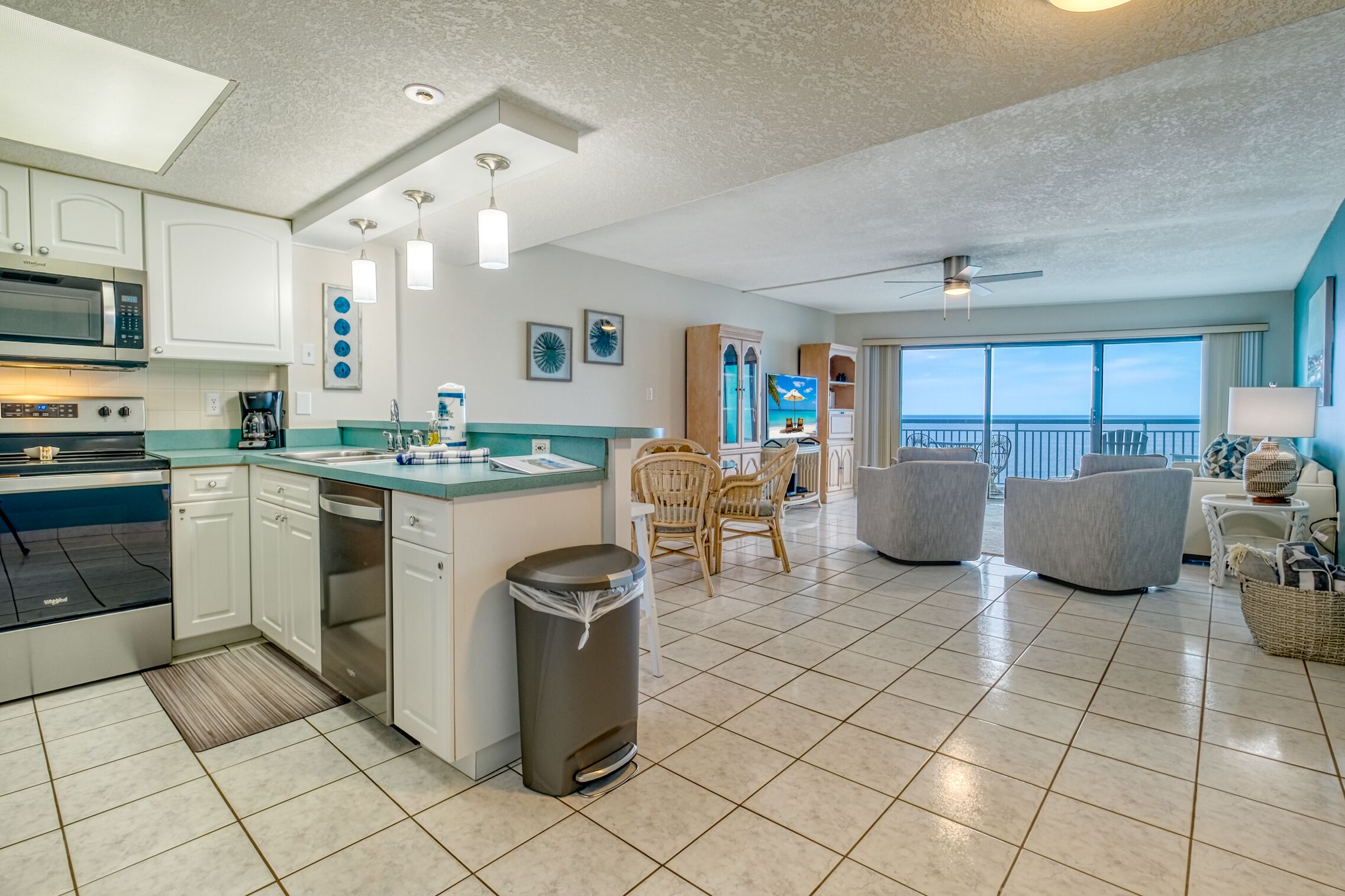 A modern kitchen with a central island, a dining area with wicker chairs, and a view of the ocean through large windows.
