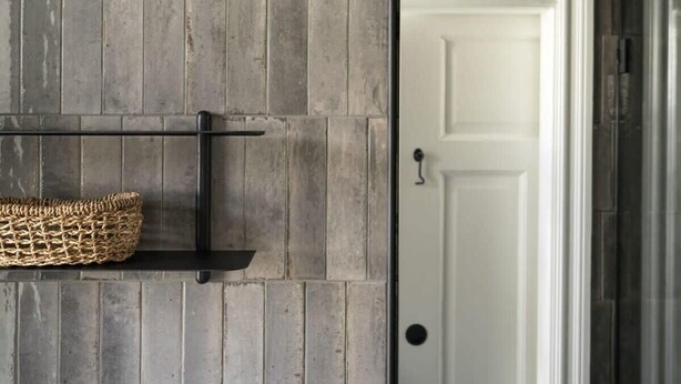 A room with a grey brick wall, a black metal shelf with a woven basket, and a white door.