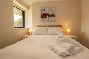 Una camera d'albergo con un letto, due abat-jour, un quadro appeso al muro e una finestra.