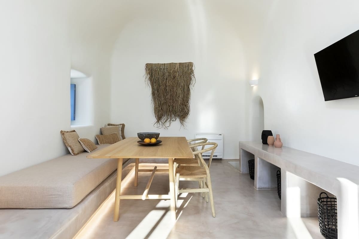 A modern, minimalist dining area with a wooden table, chairs, and a bench. A woven wall hanging decorates the space.