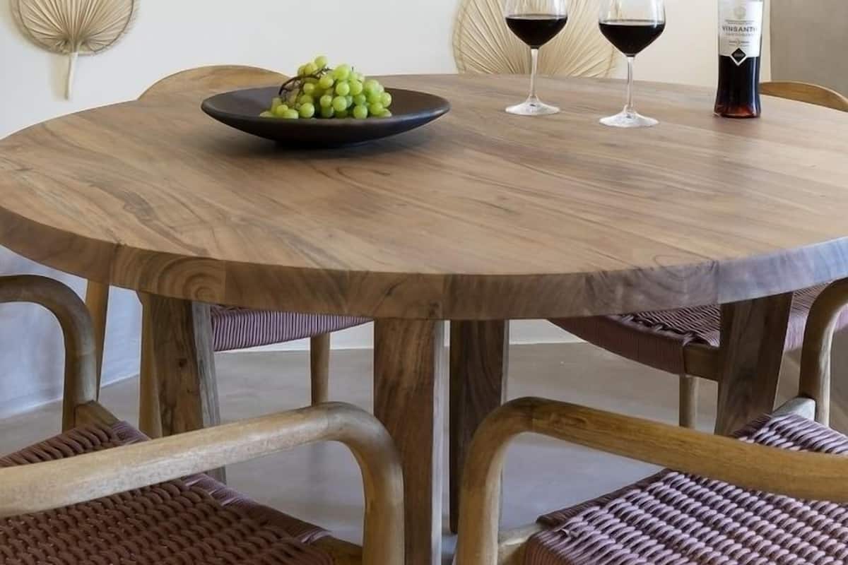 A round wooden table with a bowl of green grapes and two wine glasses, accompanied by a bottle of wine, set against a wall with decorative leaf-shaped wall art.