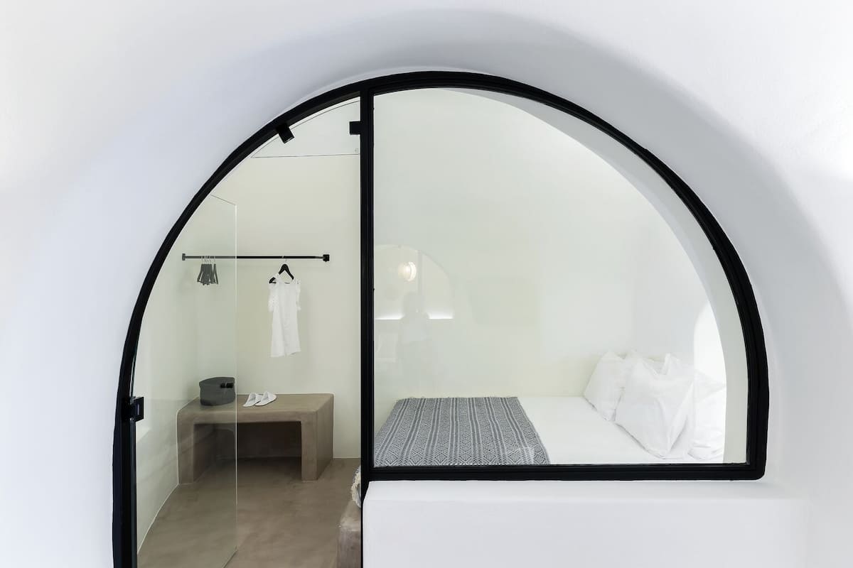 A modern, minimalist room with a curved ceiling, a bed with white bedding, a small wooden bench, and a clothing rack.
