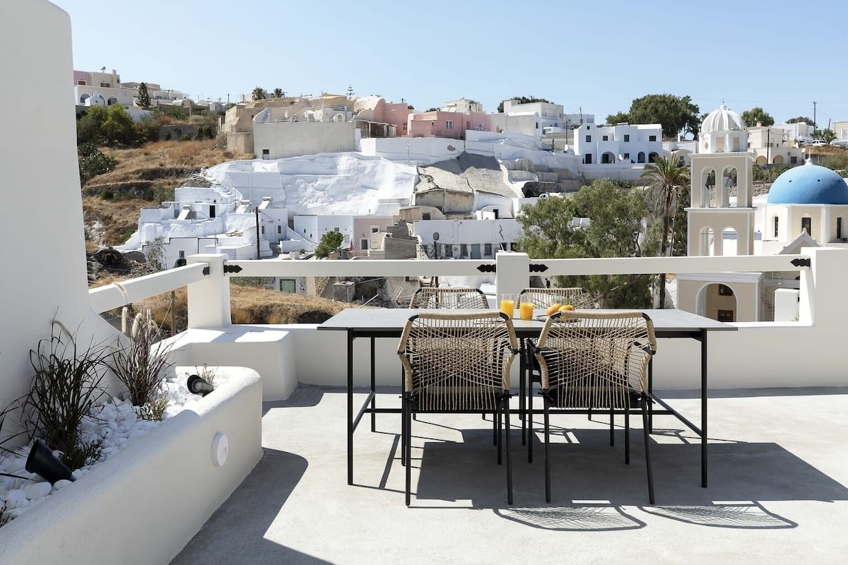 A terrace with a table and chairs overlooking a coastal town with white buildings and a blue dome.