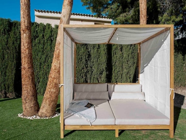 A wooden outdoor seating area with a canopy, surrounded by trees and a hedge.