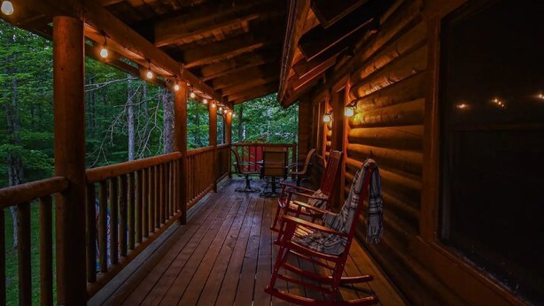 A wooden porch with string lights, rocking chairs, and a view of the forest. - Log Cabin Retreat w/ Hot Tub, Game Room, Fire Pit (Swanton)