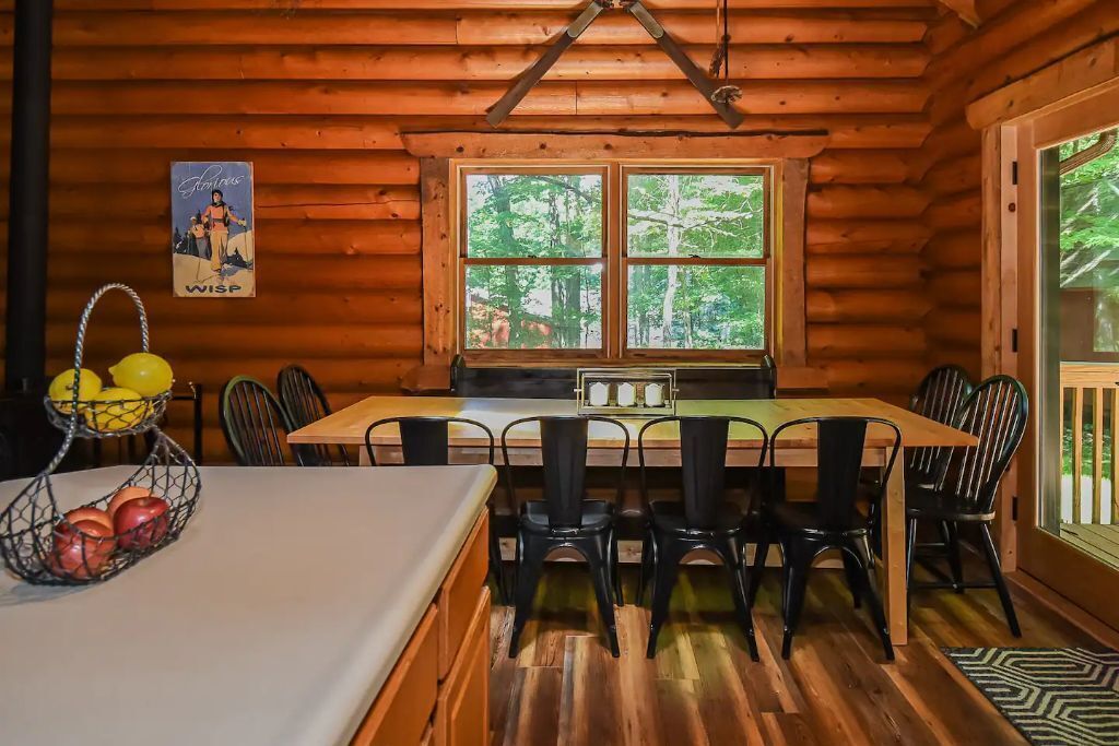 Interno di una baita in legno con un tavolo da pranzo, sedie, una finestra vista alberi e un poster appeso al muro.