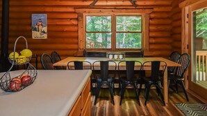 A wooden cabin interior with a dining table, chairs, a window with a view of trees, and a poster on the wall. - Log Cabin Retreat w/ Hot Tub, Game Room, Fire Pit (Swanton)