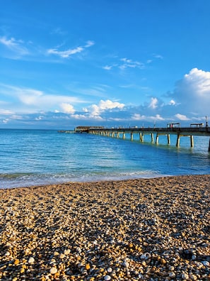 A pebbled beach with a pier extending into calm waters under a clear sky.