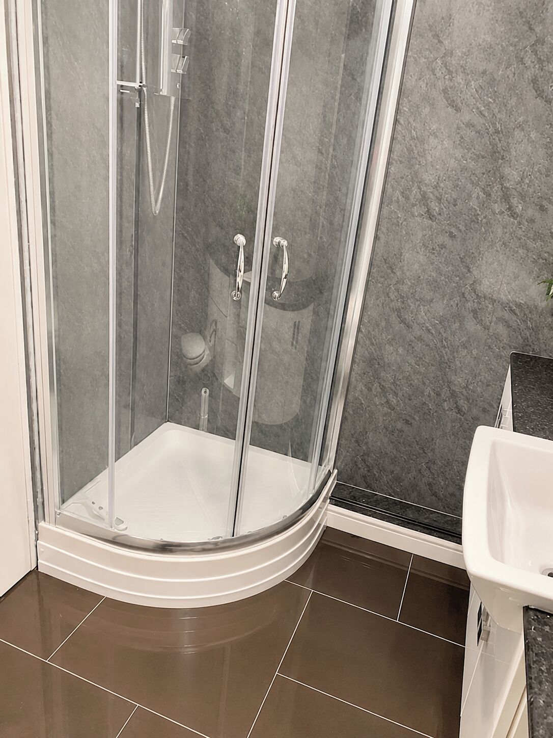 A corner shower enclosure with a glass door, a white sink, and a marble-tiled floor.