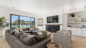 En moderne stue med en stor grå sofa, en grå lænestol og et lille rundt bord. Der er en pejs med et fladskærms-tv over, og et stort vindue med udsigt til en bygning og en swimmingpool.