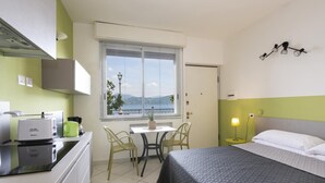 A compact hotel room with a bed, a small desk with a chair, a kitchenette, and a window with blinds. - Studio 2- lake view - Studio Apartment, Sleeps 2 (Oggebbio)