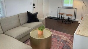 Un soggiorno con un divano beige, un tavolino rotondo con una pianta, un tappeto decorato, un tavolo in vetro con degli sgabelli e una porta con finestra.
