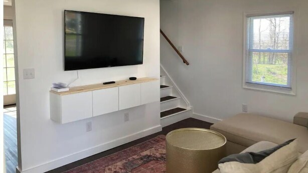 Un soggiorno moderno con una TV a schermo piatto montata su una parete bianca, una scala e un'ampia finestra con persiane.