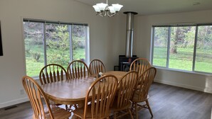 A dining room with a round wooden table and chairs, large windows with a view of greenery, a chimney, and a hanging light fixture. - Hayden’s Green Acres on Big Fall Creek, 3BR 2Bath including River Access (Fall Creek)