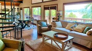 A spacious living room with a high ceiling, a staircase, and a large window offering a view of the outdoors.