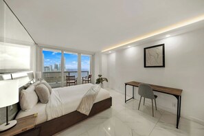 Une chambre moderne avec un lit, un bureau et une chaise, de grandes fenêtres donnant sur la ville et une œuvre d’art encadrée accrochée au mur.