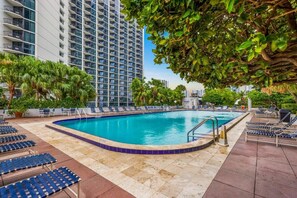 A rooftop pool with lounge chairs, surrounded by palm trees and a high-rise building.