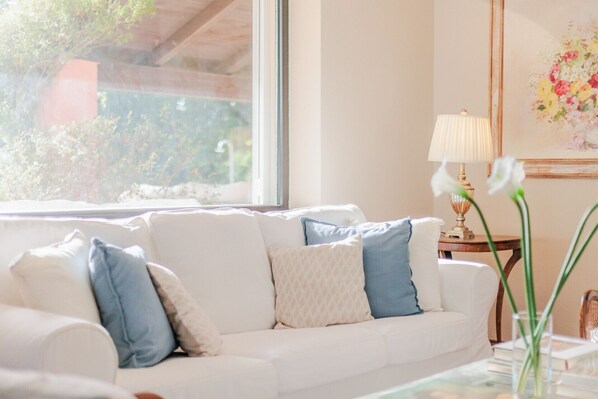 A white sofa with blue and beige cushions, a table with a lamp, and a window with a view of greenery.
