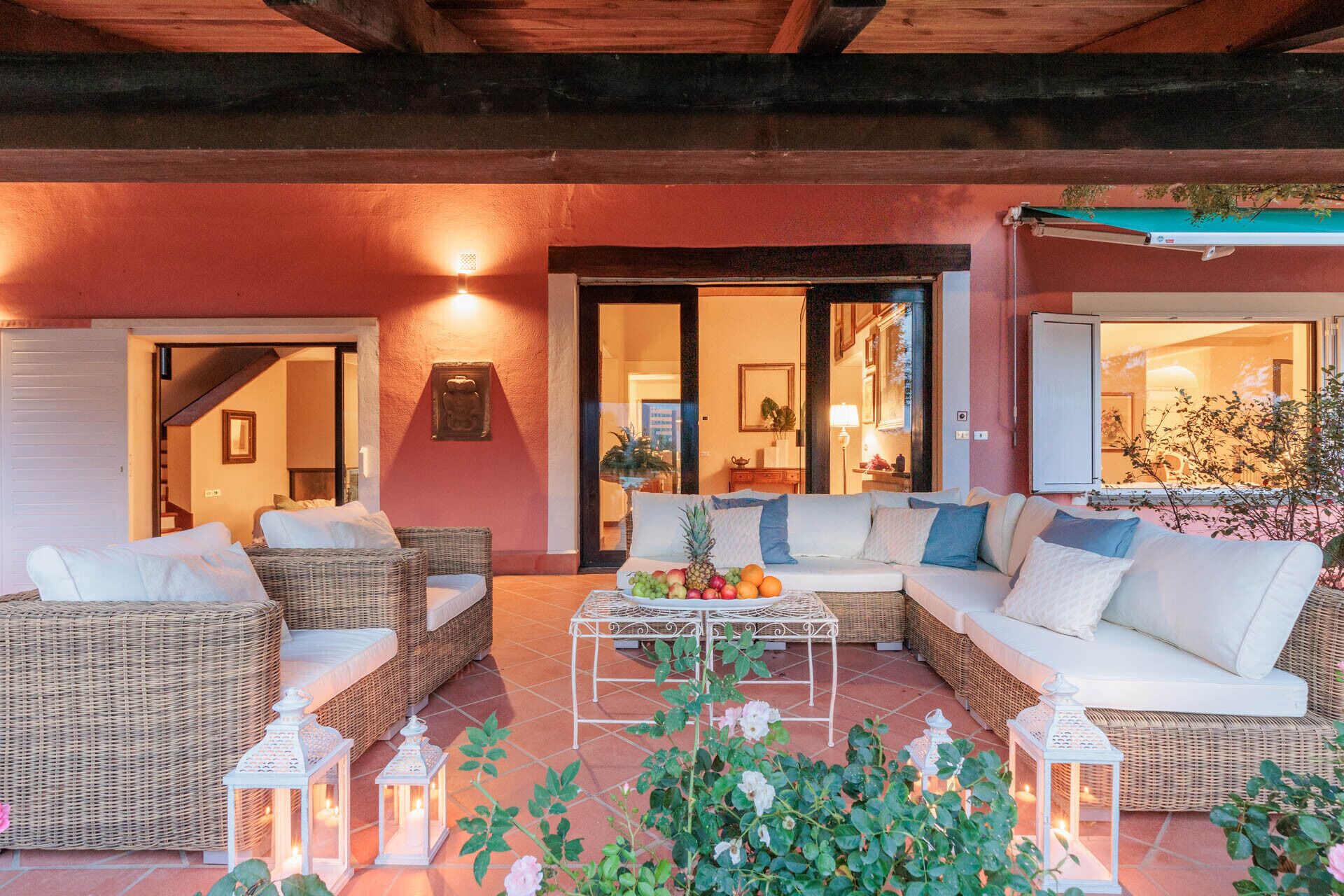 A cozy outdoor seating area with wicker furniture and white cushions, a fruit bowl on a table, and white lanterns.