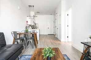 Een moderne woonkamer met een leren zwarte bank, een houten salontafel en een eethoek met metalen stoelen en een kleine plant.
