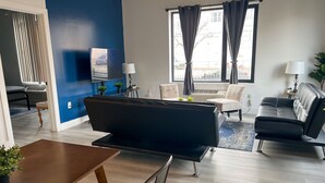 A modern living room with a dark sofa, a wooden coffee table, and a TV mounted on the wall. There are two windows with curtains, a small side table with a lamp, and a potted plant. - Cozy 2BR Near Airport 4wef (Elizabeth)