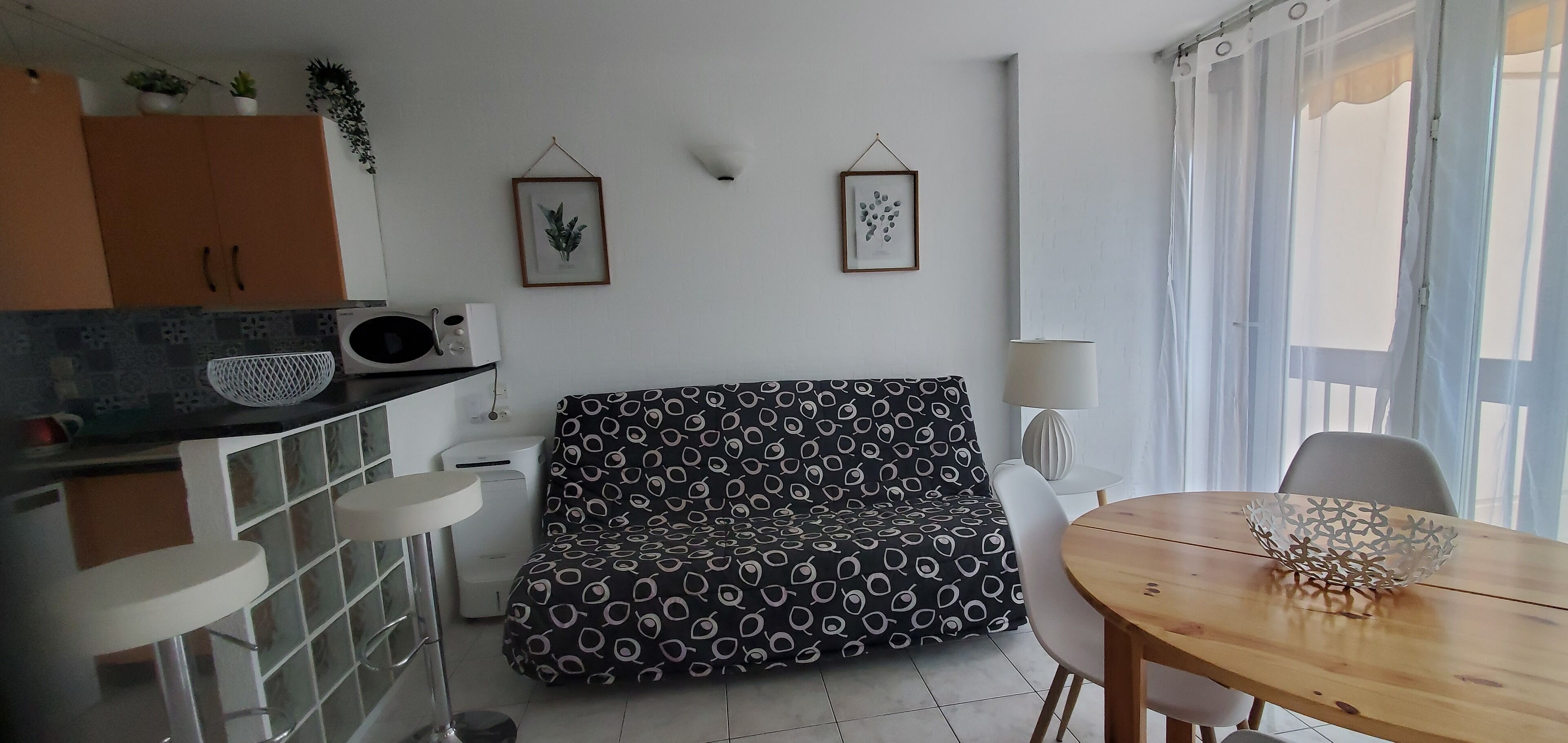 A compact living space with a kitchenette, a patterned sofa, a dining table with a decorative bowl, and a window with sheer curtains.